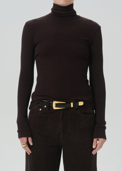 A person wearing a dark brown ribbed turtleneck sweater, dark brown pants, and a black belt with a gold buckle, shown from the neck down against a grey background.