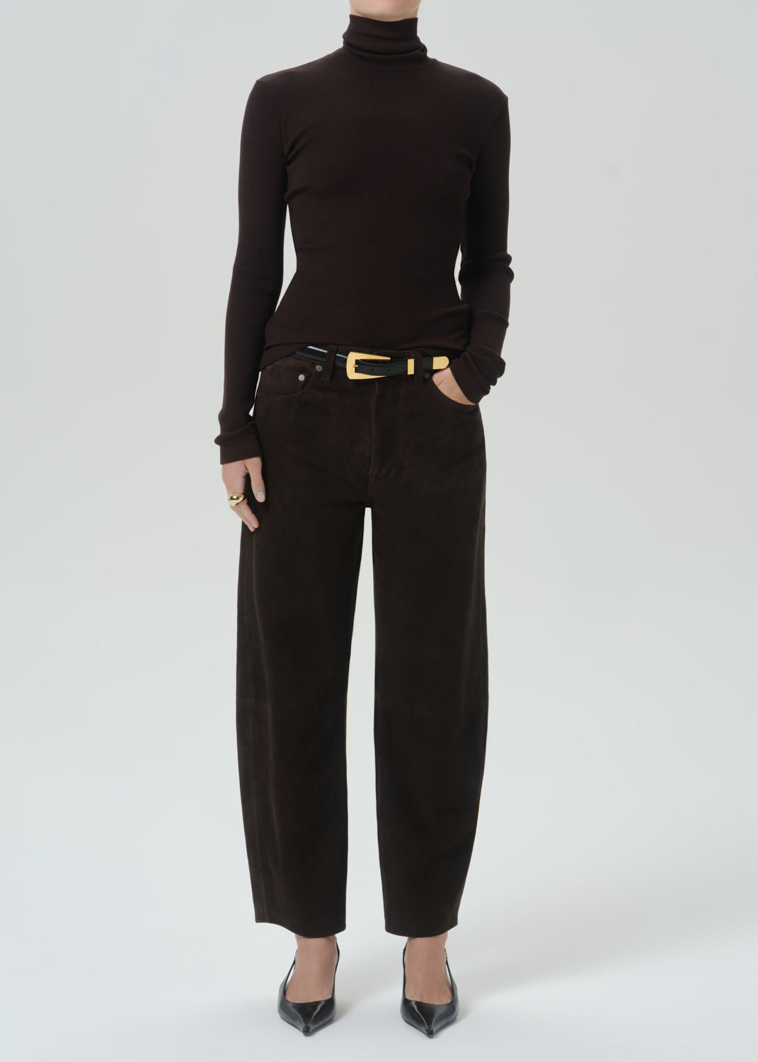 A model wearing a dark brown long-sleeve turtleneck sweater and matching dark brown wide-leg trousers with a black belt featuring a gold buckle, paired with black pointed-toe heels, against a grey background.