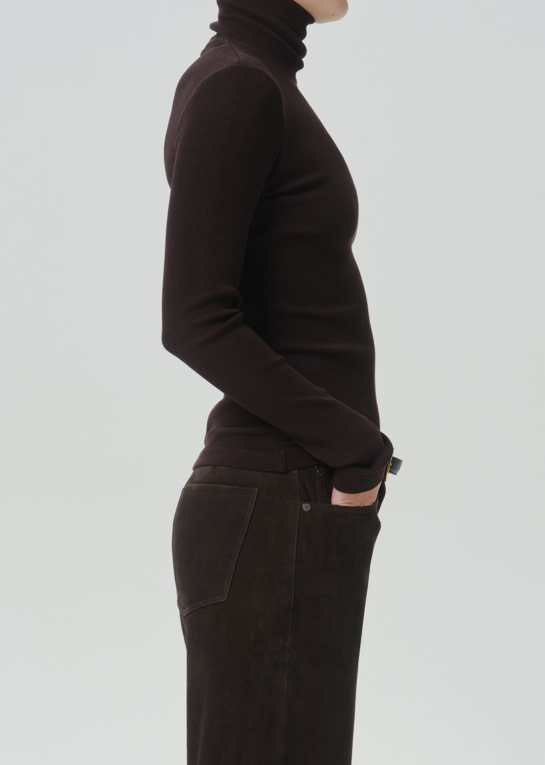 Side profile of a model wearing a dark brown ribbed turtleneck sweater and matching dark brown corduroy trousers against a grey background.