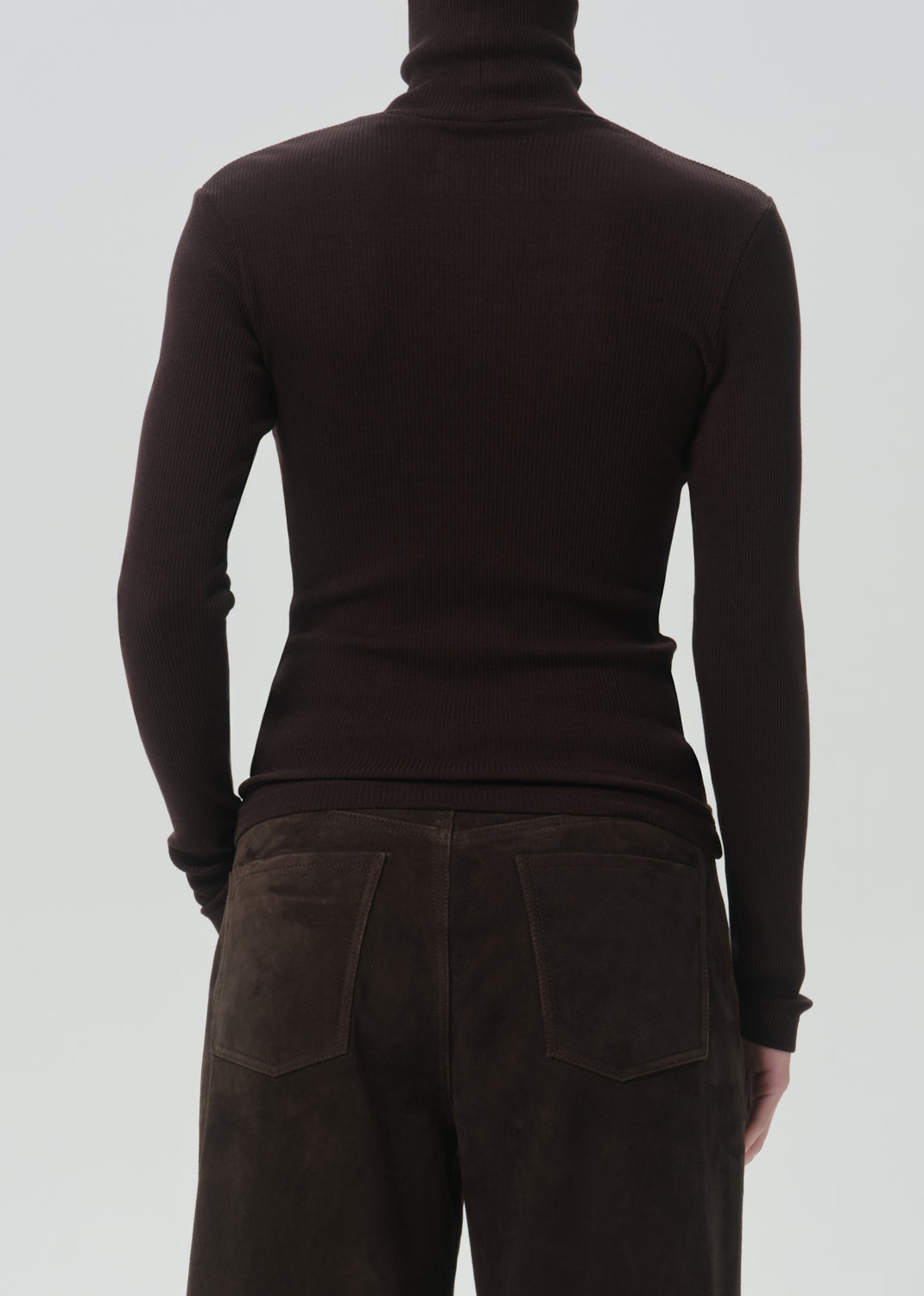 Rear view of a person wearing a dark brown ribbed turtleneck sweater and dark brown pants against a grey background.