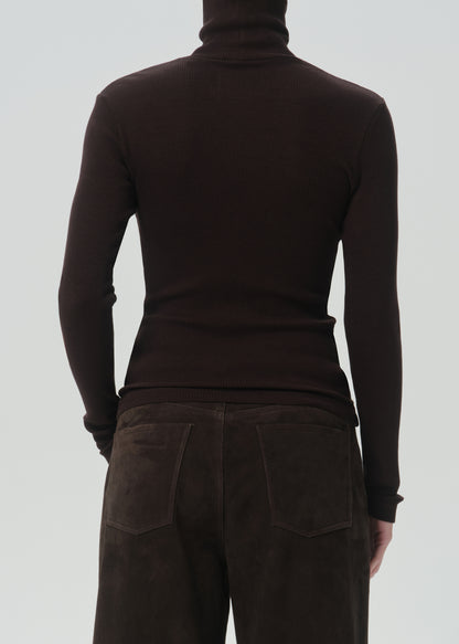 Rear view of a person wearing a dark brown ribbed turtleneck sweater and dark brown pants against a grey background.