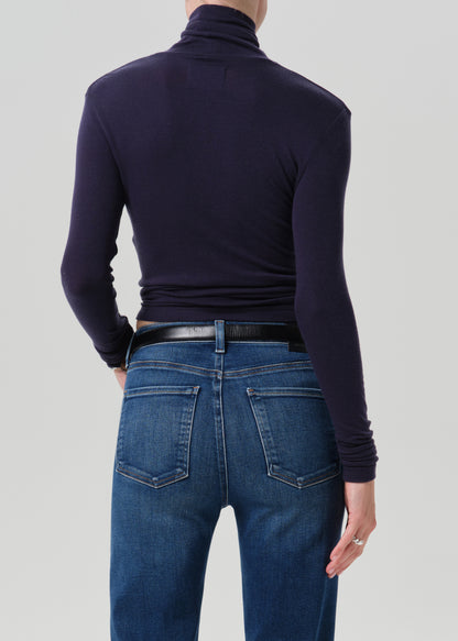 Person wearing a navy long sleeve turtleneck with dark wash straight leg jeans viewed from the back on a grey background