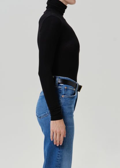 Woman wearing a black turtleneck top with medium blue straight leg jeans viewed from the side on a grey background.