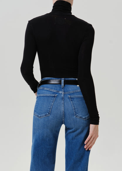 Woman wearing a black turtleneck top with medium blue straight leg jeans viewed from the back on a grey background.