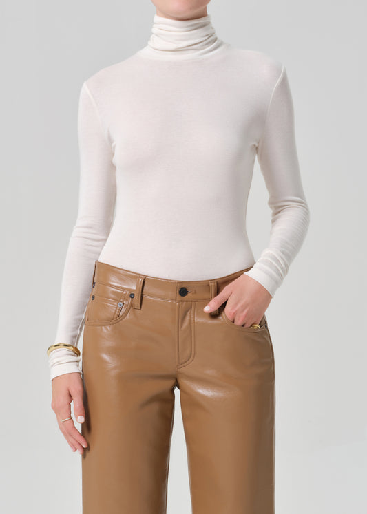 Person wearing a white turtleneck and brown leather pants on a gray background