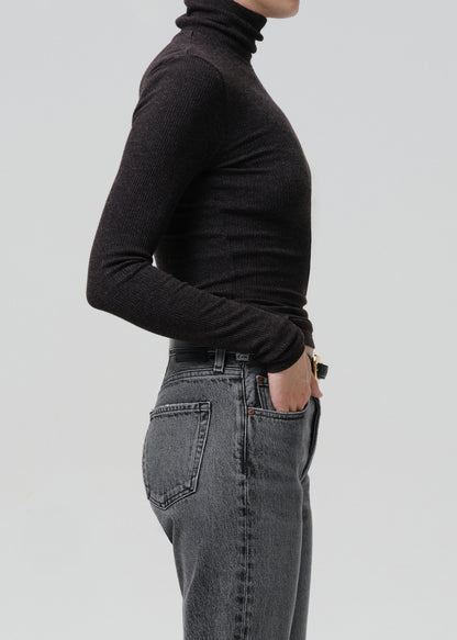 Person wearing a black ribbed long sleeve turtleneck with grey straight leg jeans viewed from the side on a grey background
