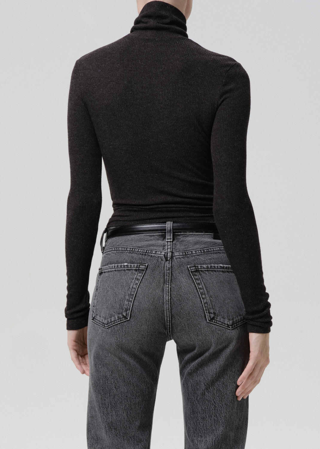 Person wearing a black ribbed long sleeve turtleneck with grey straight leg jeans viewed from the back on a grey background