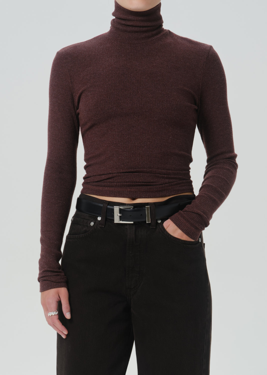 A model wearing a dark burgundy ribbed long-sleeve turtleneck top paired with black jeans and a black belt, shown from the neck down against a grey background.