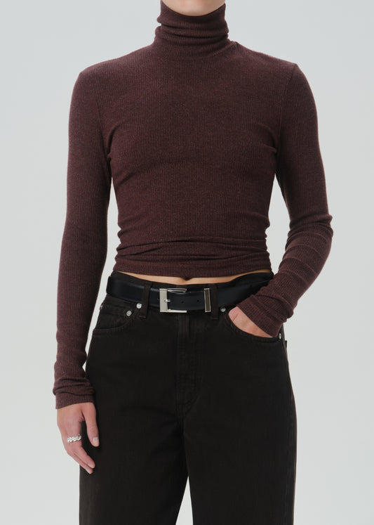 A model wearing a dark burgundy ribbed long-sleeve turtleneck top paired with black jeans and a black belt, shown from the neck down against a grey background.