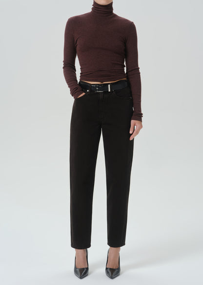 A model wearing a dark brown ribbed turtleneck top, black high-waisted cropped trousers with a black belt, and black pointed-toe heels, photographed against a grey background.