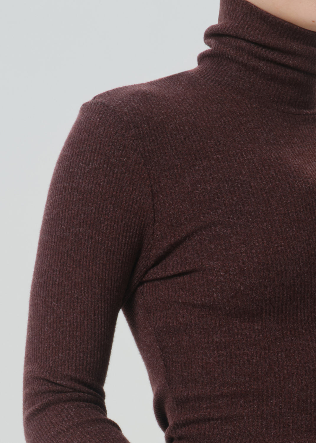 Close-up of a person wearing a ribbed long-sleeve turtleneck sweater in a deep burgundy-brown color, against a grey background.