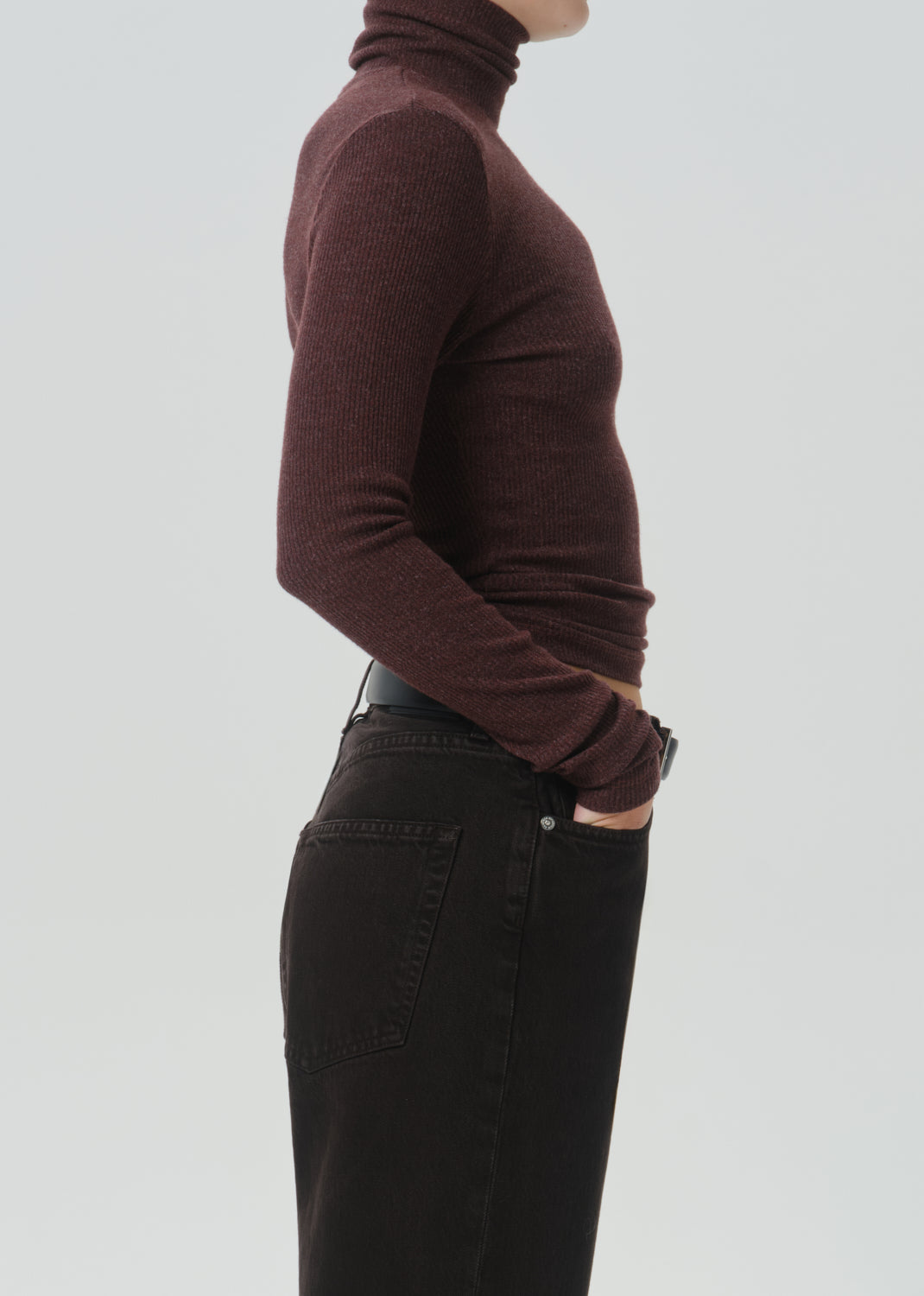 A side-profile view of a person wearing a ribbed, long-sleeve turtleneck sweater in a deep burgundy color, paired with black jeans, against a grey background.