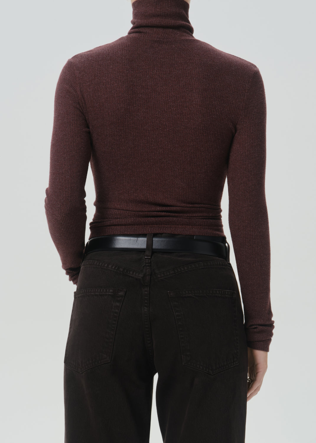Rear view of a person wearing a fitted ribbed turtleneck sweater in deep burgundy, paired with black pants and a black belt, against a grey background.
