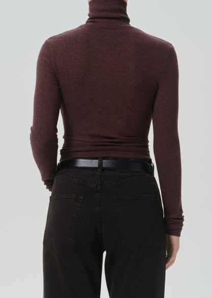 Rear view of a person wearing a fitted ribbed turtleneck sweater in deep burgundy, paired with black pants and a black belt, against a grey background.
