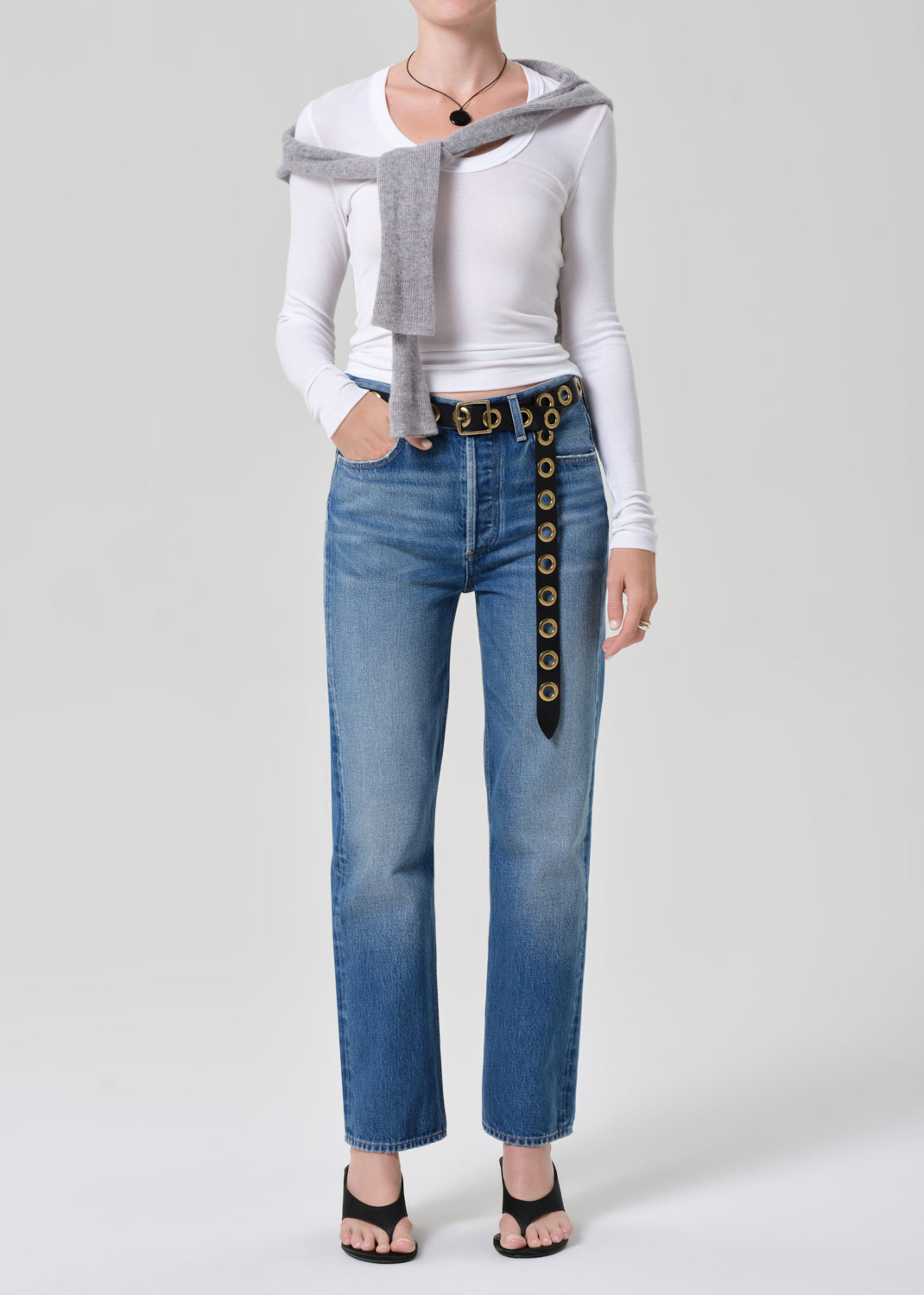 Person wearing a white long-sleeve shirt, gray sweater, and blue jeans with a decorative belt on a light gray background.