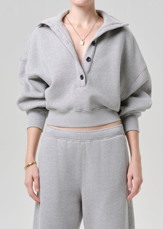 A model wearing a light grey cropped hoodie sweatshirt with a buttoned collar and matching light grey sweatpants, shown from the neck down against a neutral light grey background.