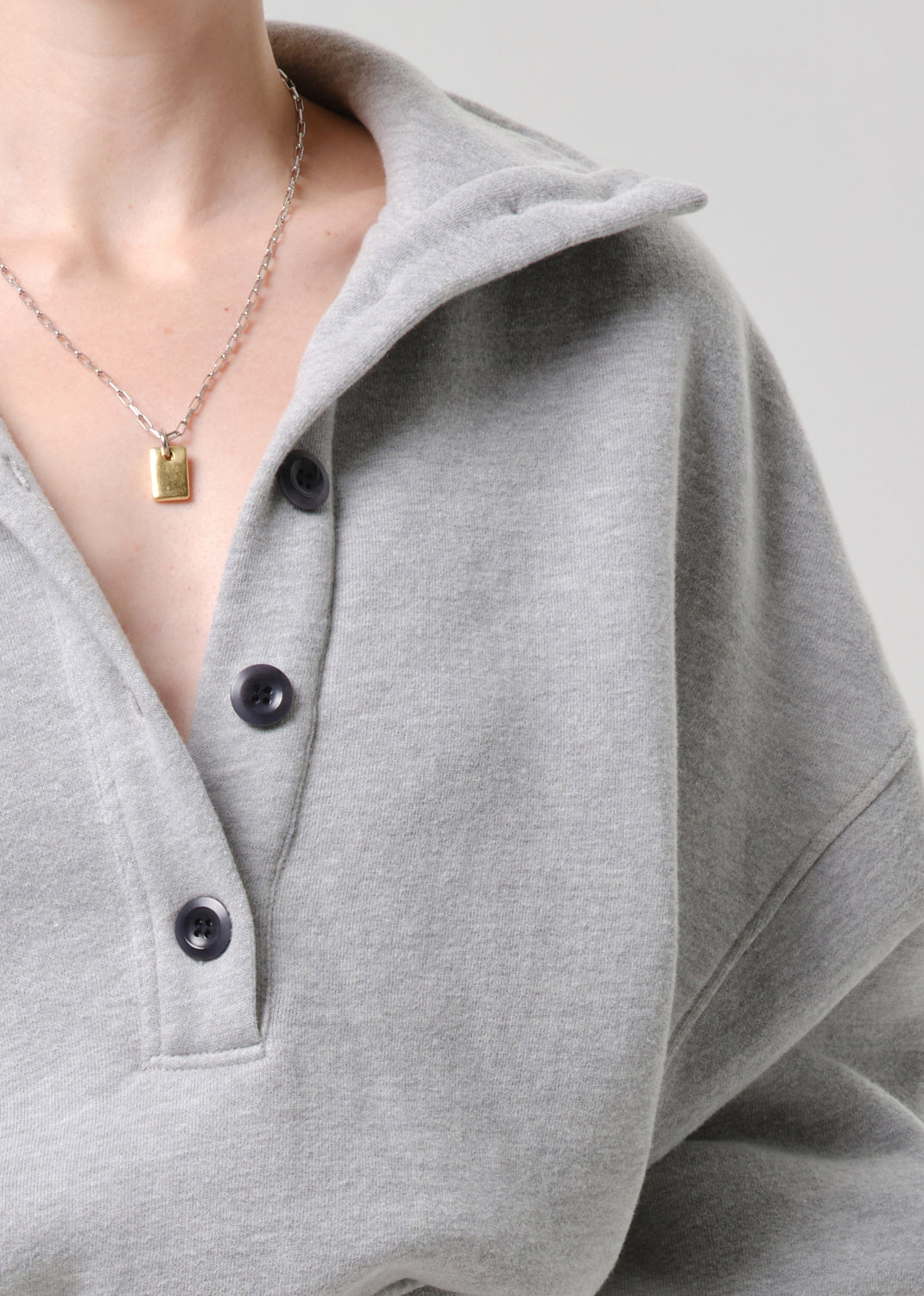 Close-up of a person wearing a light grey heathered pullover top with a partial stand collar and dark buttons, paired with a silver chain necklace featuring a small gold rectangular pendant.