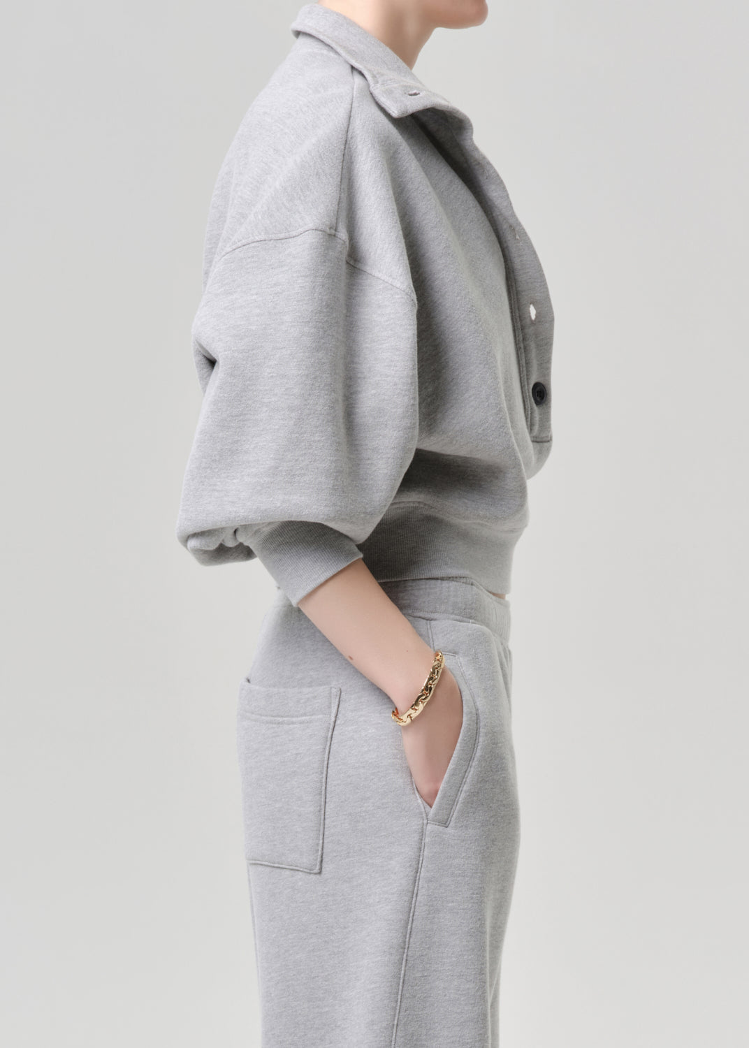 A side-profile view of a model wearing a light grey heathered matching lounge set, featuring a cropped button-front sweatshirt and high-waisted sweatpants with side pockets, accessorized with a gold chain bracelet.