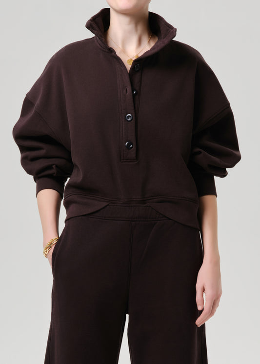 A model wearing a dark brown pullover sweatshirt with a high mock neck and button placket, paired with matching dark brown sweatpants, against a grey background.