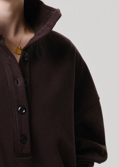 Close-up of a person wearing a dark brown button-front top or sweatshirt with a high collar and black buttons, against a grey background.