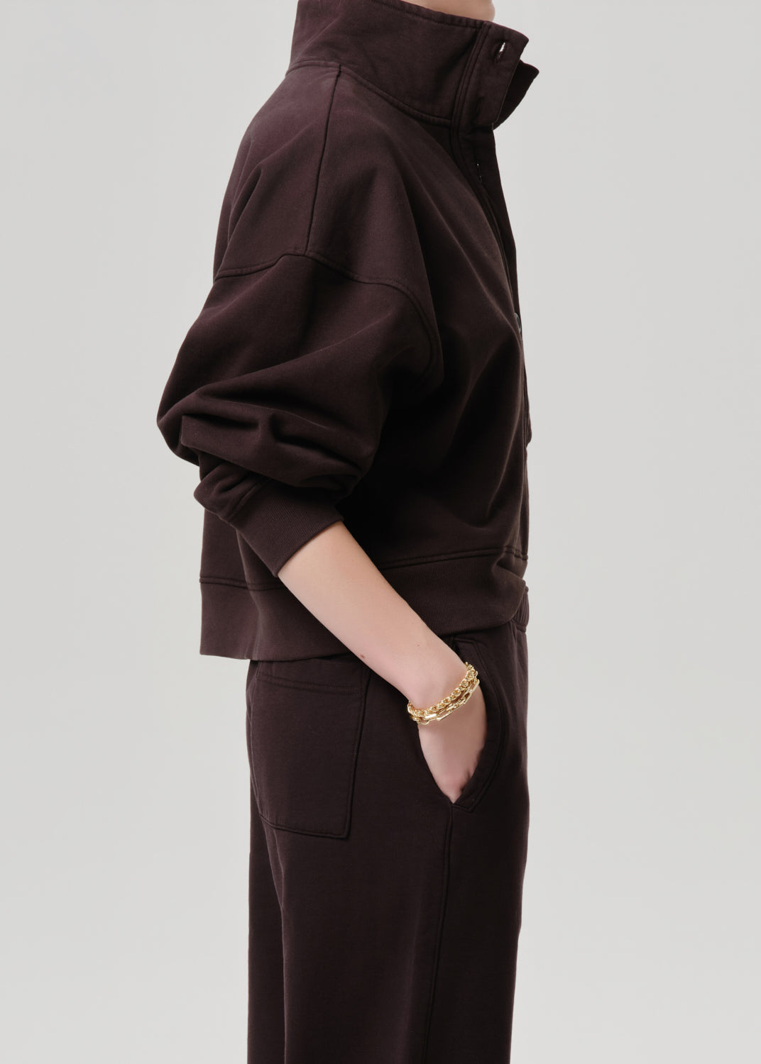 A person shown in side profile wearing a dark brown cropped zip-up sweatshirt and matching dark brown pants, with a gold chain bracelet visible on one wrist, against a grey background.