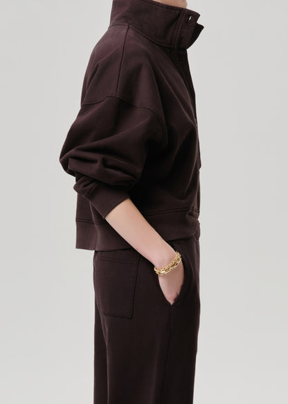 A person shown in side profile wearing a dark brown cropped zip-up sweatshirt and matching dark brown pants, with a gold chain bracelet visible on one wrist, against a grey background.