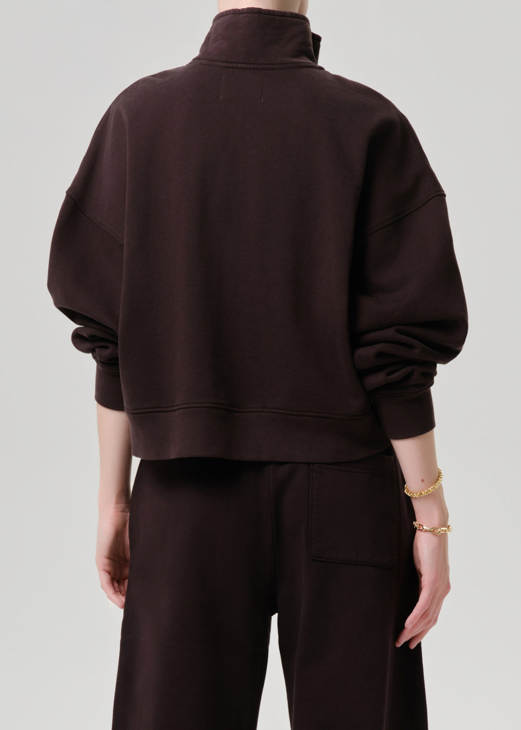 A person shown from the back wearing an oversized dark brown mock-neck sweatshirt and matching dark brown pants against a grey background.