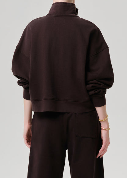 A person shown from the back wearing an oversized dark brown mock-neck sweatshirt and matching dark brown pants against a grey background.
