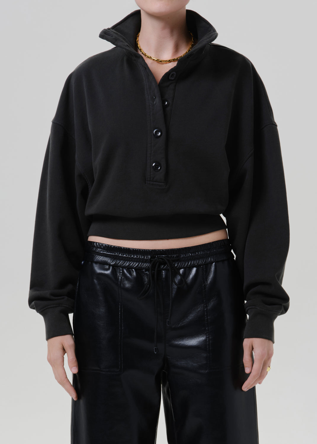 Person wearing a black long sleeve collared pullover with a partial button placket, black leather pants, and a gold necklace, front cropped view on a grey background.