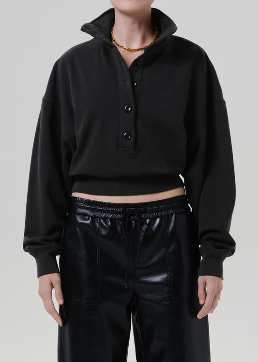 Person wearing a black long sleeve collared pullover with a partial button placket, black leather pants, and a gold necklace, front cropped view on a grey background.
