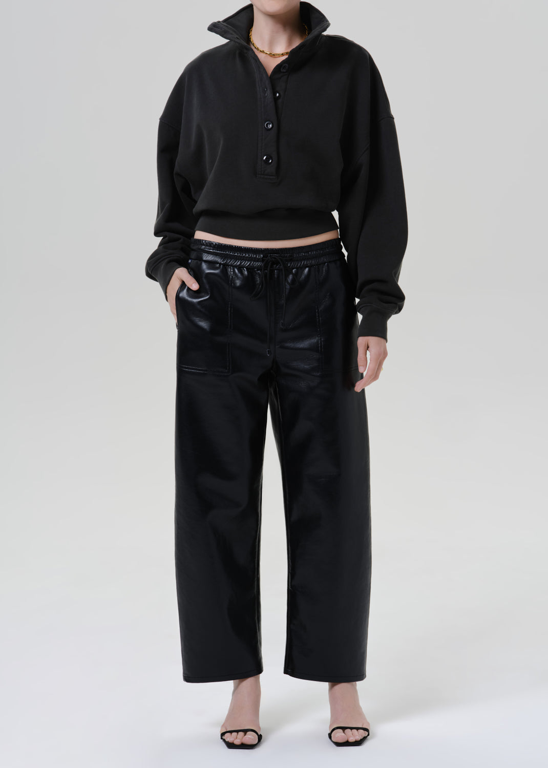 Person wearing a black long sleeve collared pullover with black leather pants and black heels, front full body view on a grey background.