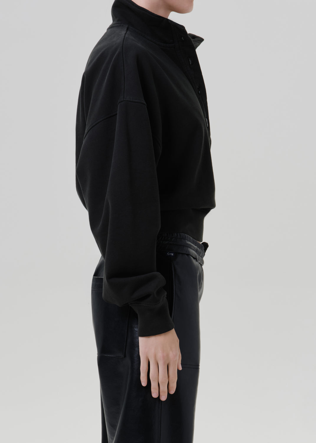 Person wearing a black long sleeve collared pullover with black leather pants viewed from the side on a grey background