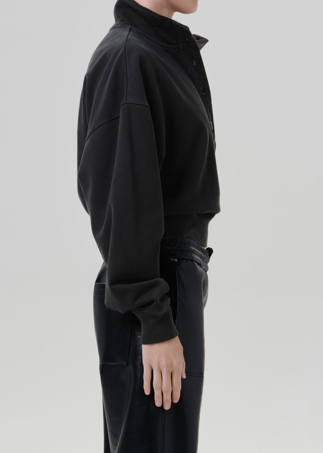 Person wearing a black long sleeve collared pullover with black leather pants, side view on a grey background.