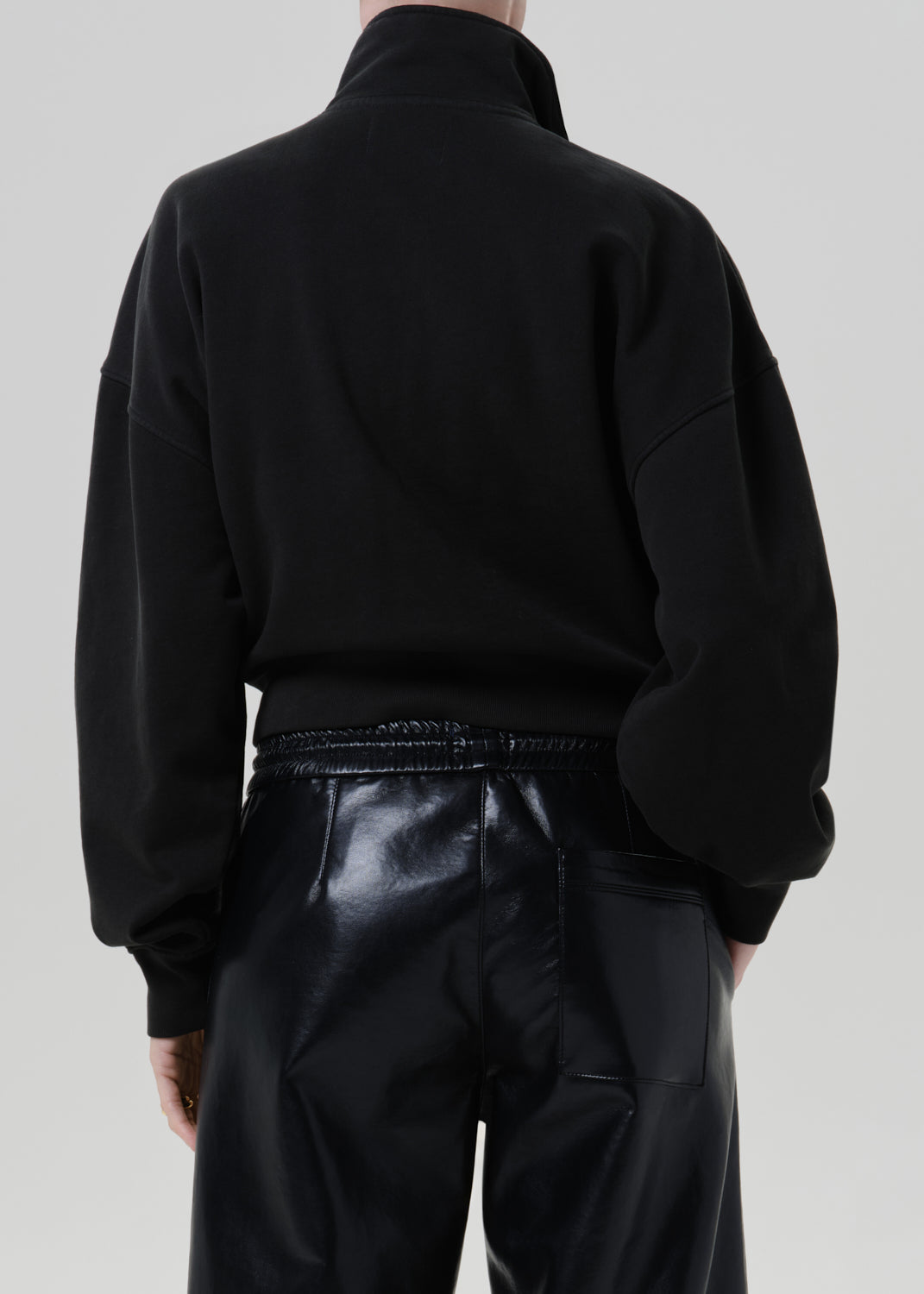 Person wearing a black long sleeve collared pullover with black leather pants viewed from the back on a grey background