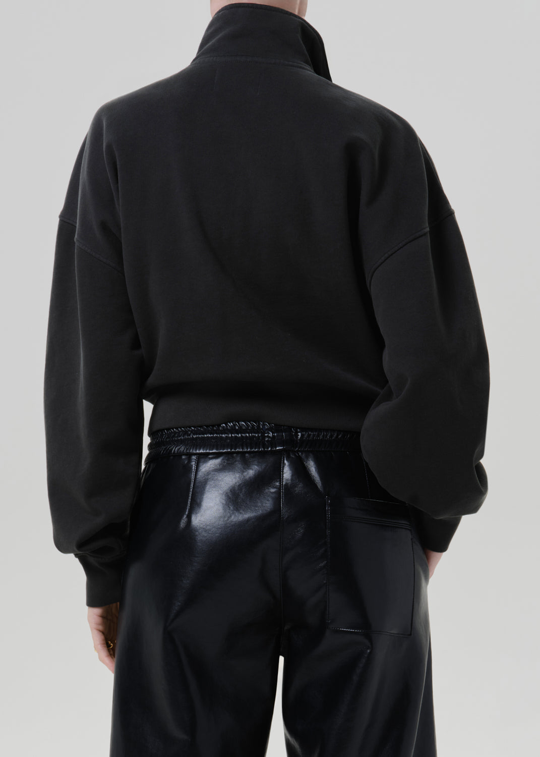 Person wearing a black long sleeve collared pullover with black leather pants, back cropped view on a grey background.