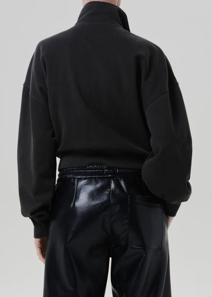 Person wearing a black long sleeve collared pullover with black leather pants, back cropped view on a grey background.