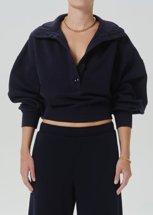 A model wearing a dark navy blue cropped pullover hoodie with a short button placket and oversized balloon sleeves, paired with matching dark pants against a grey background.