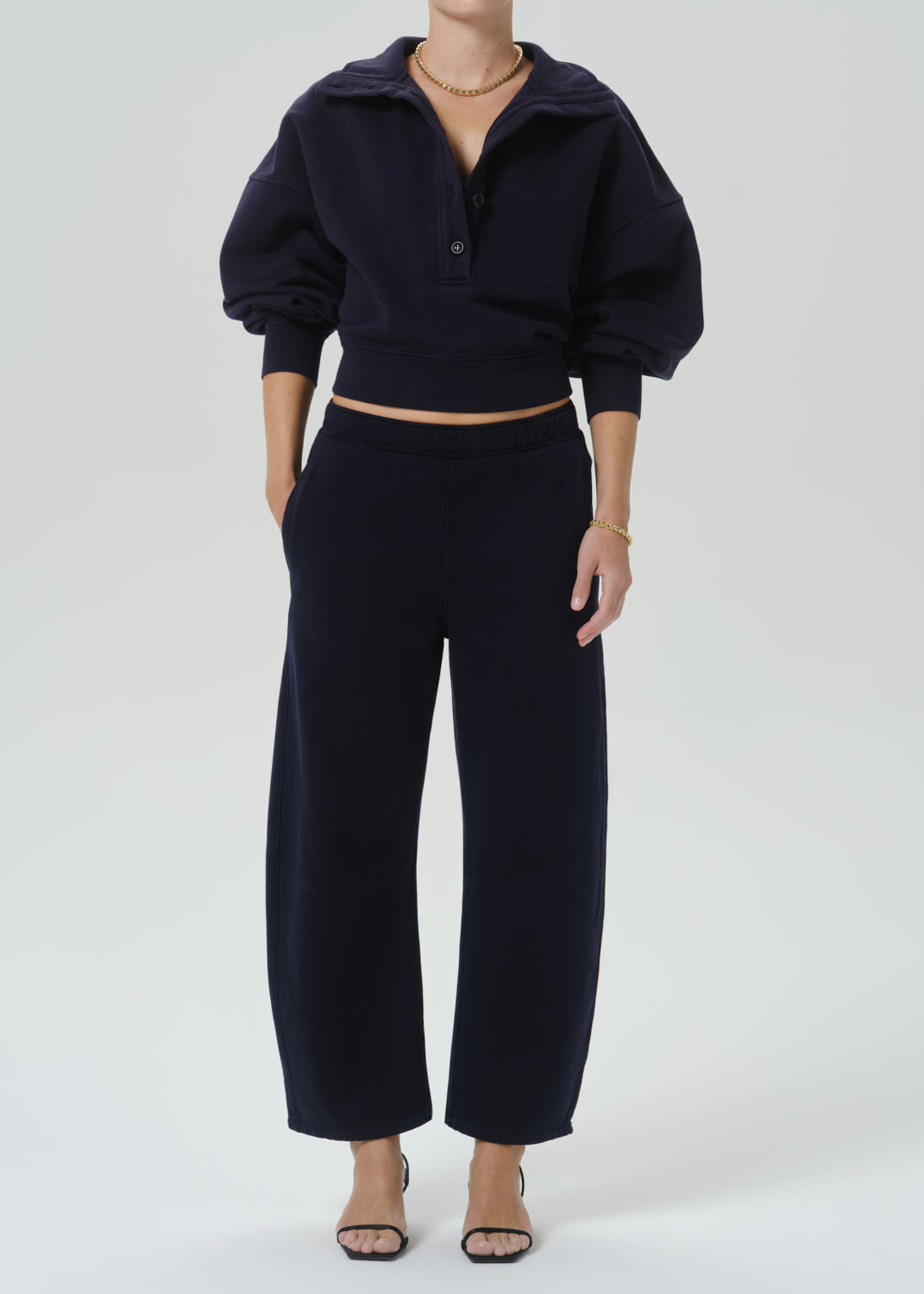 A model wearing a dark navy blue two-piece lounge set consisting of a cropped long-sleeve henley top with puffed sleeves and matching wide-leg cropped pants, styled with neutral sandals and gold jewelry against a grey background.