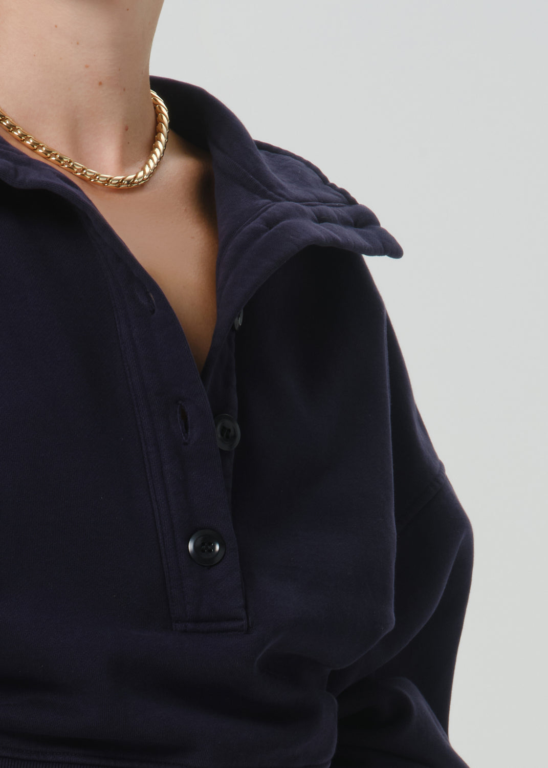 Close-up of a person wearing a dark navy long-sleeve collared top with a partial button placket and a chunky gold chain necklace, against a grey background.