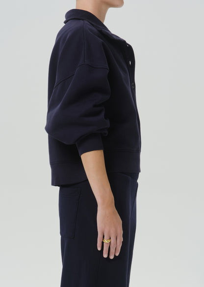 A side-profile view of a model wearing a dark navy blue cropped jacket with a high collar and matching navy pants, set against a grey background.
