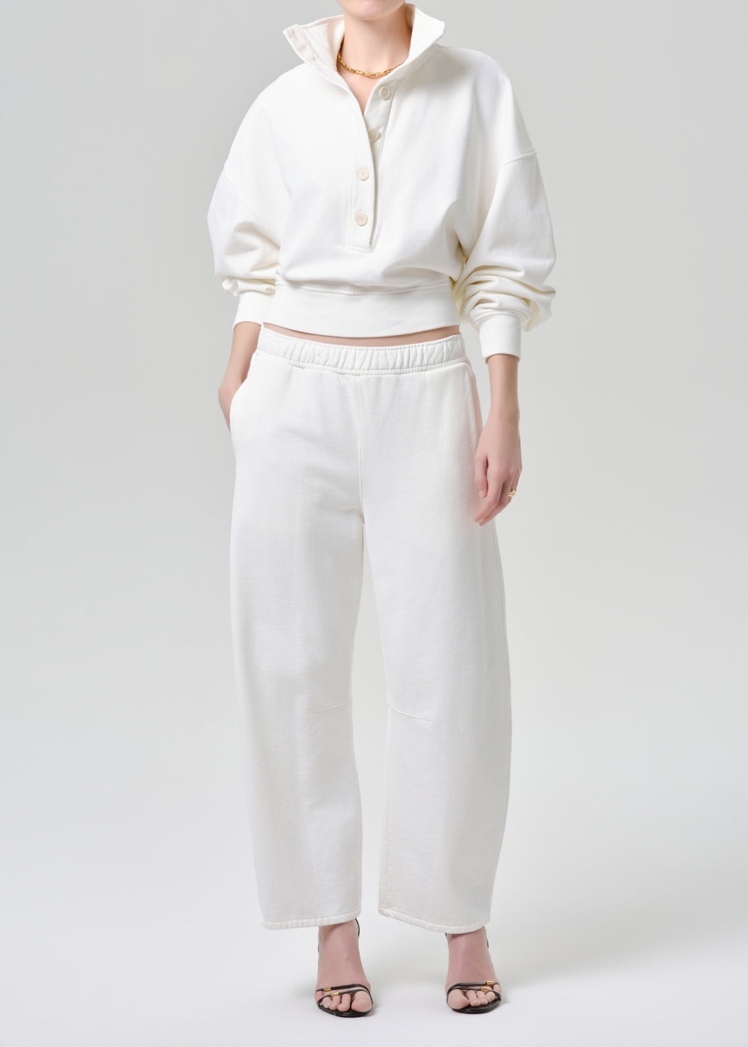 A model wearing a matching off-white two-piece lounge set consisting of a cropped long-sleeve button-up sweatshirt and wide-leg cropped pants, styled with black heeled sandals against a grey background.