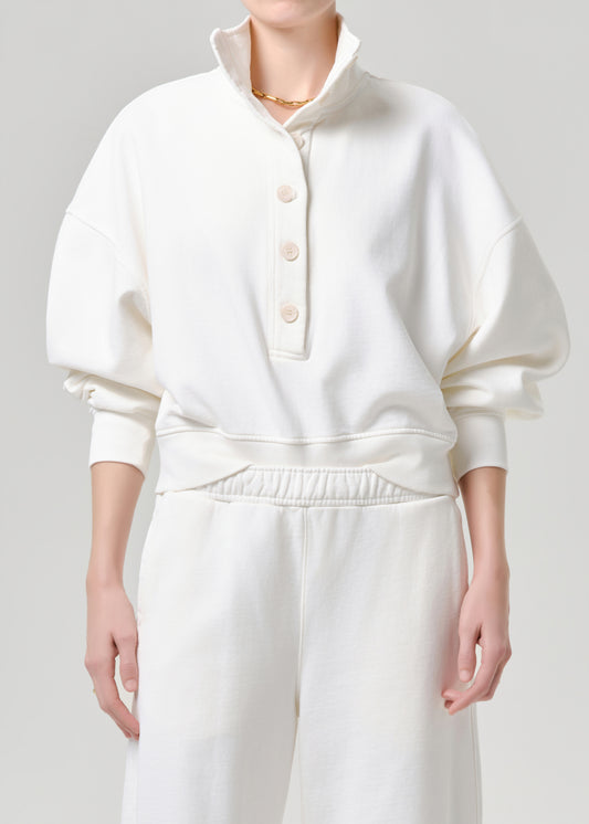 A model wearing a cream-colored pullover sweatshirt with a high mock neck, three-quarter button placket, and voluminous balloon sleeves, paired with matching cream sweatpants against a grey background.