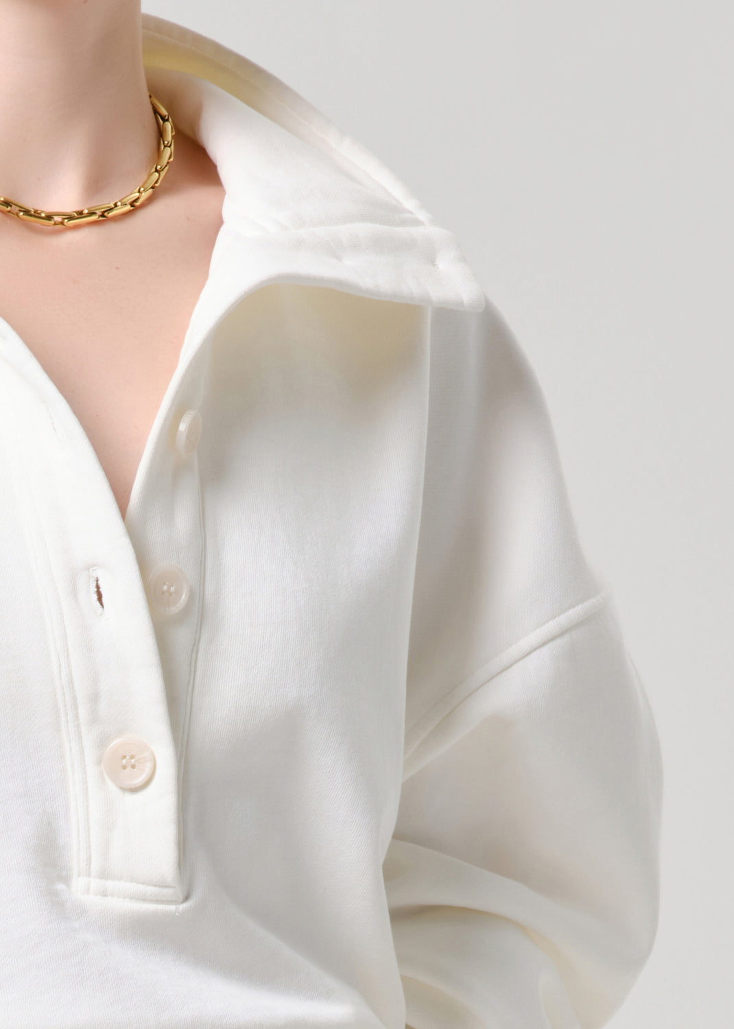 Close-up of a person wearing a white button-front collared shirt with a gold chain necklace against a grey background.