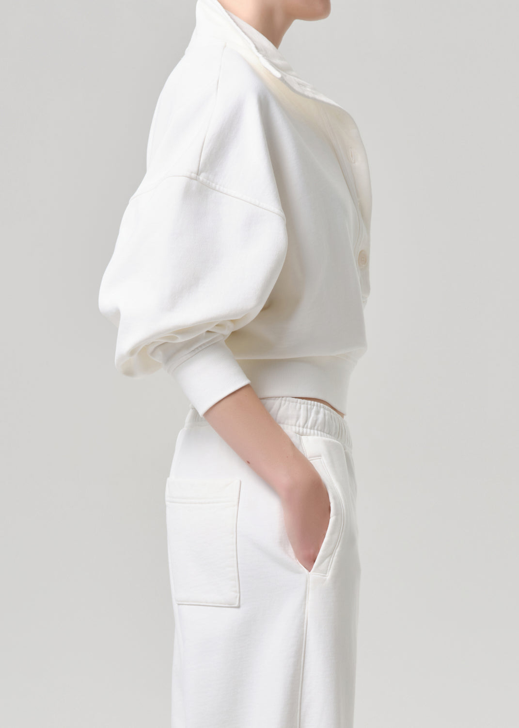 A model shown from the side wearing a white cropped sweatshirt with voluminous balloon sleeves and matching white trousers, set against a grey background.