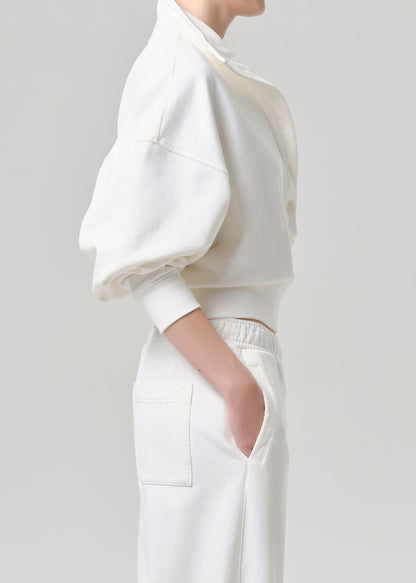 A model shown from the side wearing a white cropped sweatshirt with voluminous balloon sleeves and matching white trousers, set against a grey background.