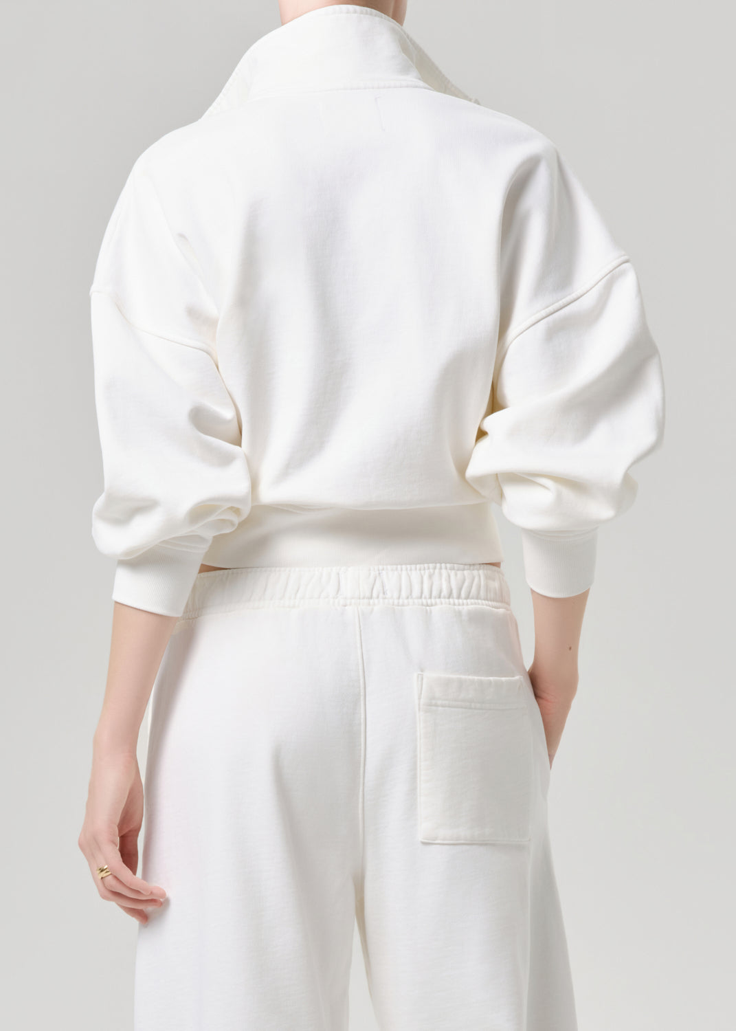 A model shown from the back wearing a white high-neck sweatshirt and matching white sweatpants against a grey background.