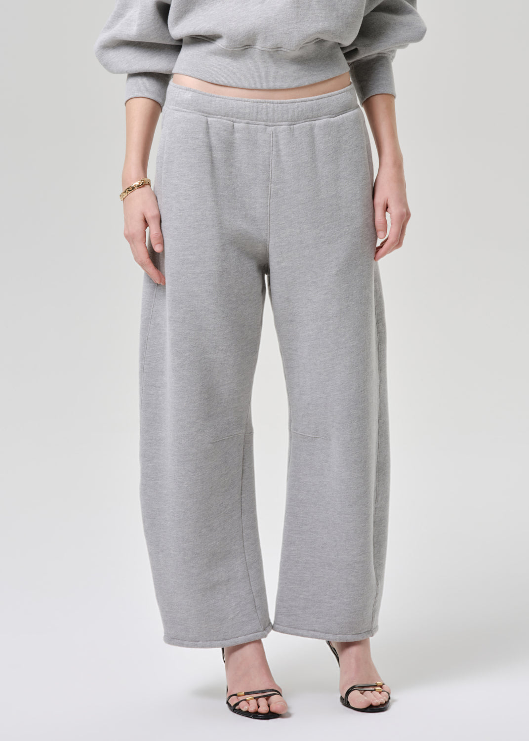 A model wearing light grey wide-leg sweatpants and a matching gray cropped sweatshirt, paired with black strappy heels, against a light grey studio background.