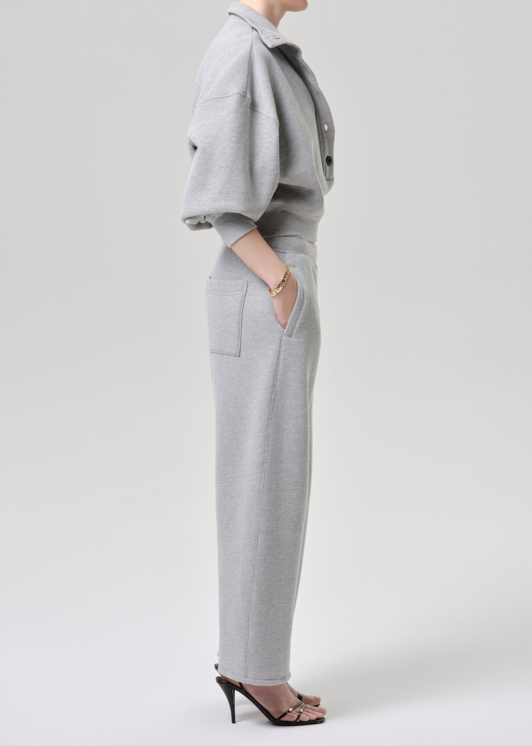 A model shown from the side wearing a light grey knit matching set consisting of a cropped, short-sleeved button-up shirt and high-waisted wide-leg trousers with side pockets, paired with black high-heeled sandals.
