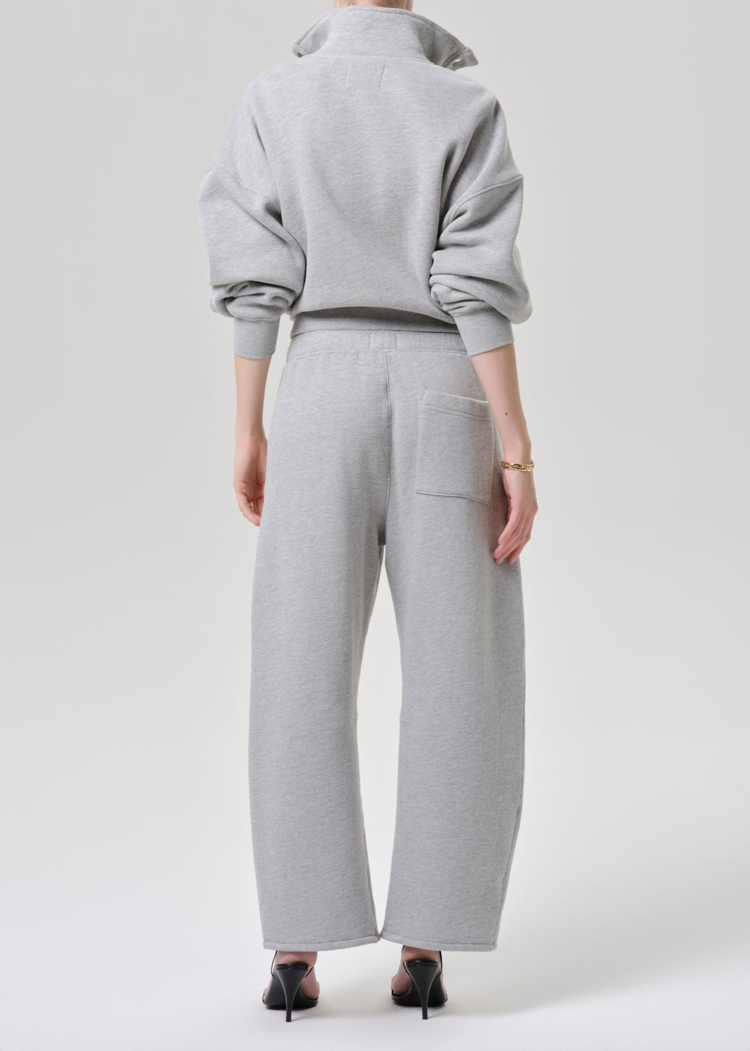 A model shown from the back wearing a light grey heathered sweatshirt and matching wide-leg sweatpants, paired with black pointed-toe heels against a grey background.