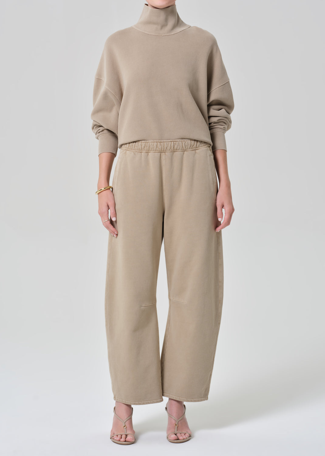 Person wearing a beige turtleneck sweater and pants on a grey background